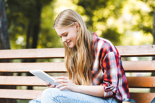 selective focus of beautiful girl in casual clothes sitting on wooden bench in park and using digital tablet