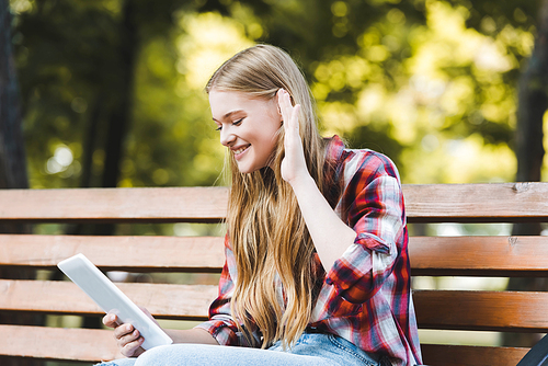 selective focus of beautiful girl in casual clothes sitting on wooden bench in park and using digital tablet and gesturing