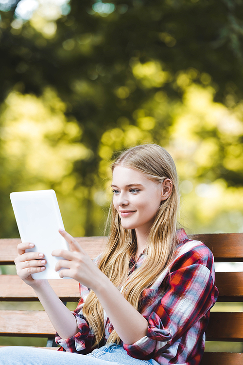 selective focus of beautiful young girl in casual clothes sitting on wooden bench in park and using digital tablet