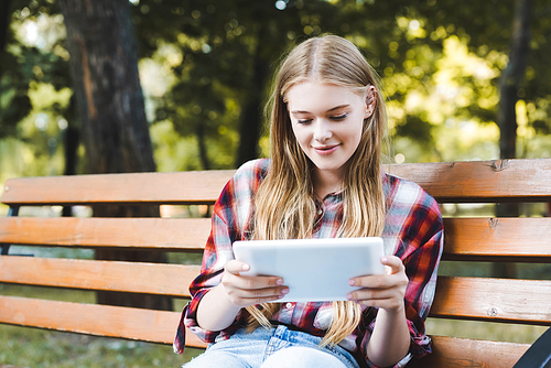 young girl in casual clothes sitting on wooden bench in park and using digital tablet