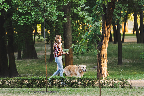 full length view of beautiful girl in casual clothes waking in park with golden retriever and talking on smartphone