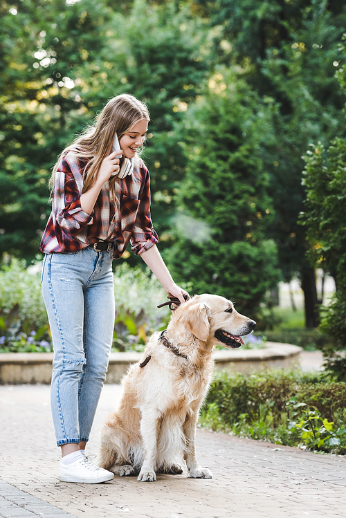 full length view of beautiful young girl in casual clothes waking in park with golden retriever and talking on smartphone