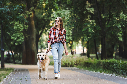 full length view of beautiful girl in casual clothes waking in park with golden retriever