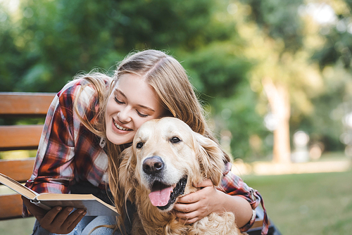 beautiful girl in casual clothes reading book and hugging golden retriever while sitting on wooden bench in park