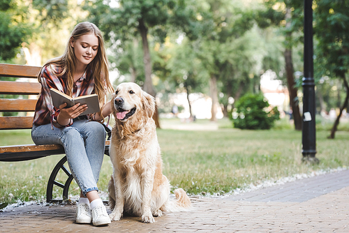 full length view of beautiful girl in casual clothes reading book and petting golden retriever while sitting on wooden bench