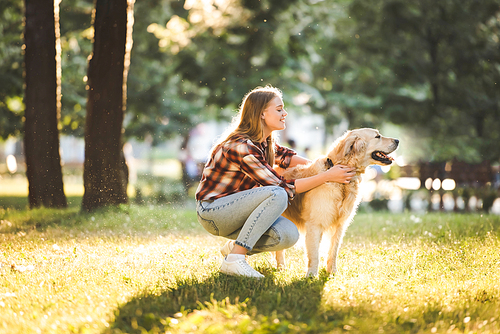 side view of beautiful girl in casual clothes petting golden retriever while sitting on meadow in sunlight