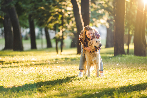 full length view of beautiful girl in casual clothes hugging golden retriever while standing on meadow in sunlight