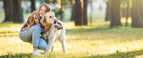 panoramic shot of beautiful young girl in casual clothes hugging golden retriever while sitting on meadow in sunlight