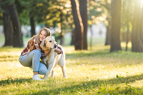full length view of beautiful young girl in casual clothes hugging golden retriever while sitting on meadow in sunlight and 