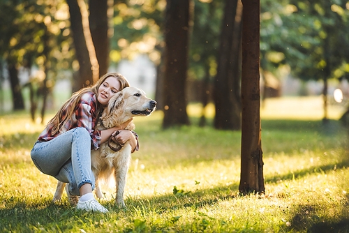 full length view of beautiful young girl in casual clothes hugging golden retriever while sitting on meadow in sunlight and looking away