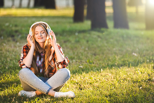 full length view of beautiful girl in casual clothes sitting with crossed legs on meadow in sunlight and listening to music on headphones