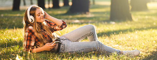 panoramic shot of beautiful girl in casual clothes lying on meadow in sunlight and listening to music on headphones