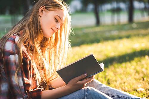 side view of beautiful girl in casual clothes sitting on meadow in sunlight and reading book