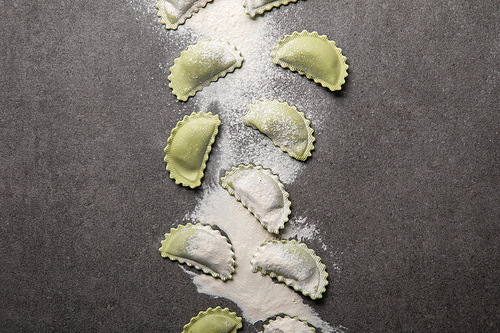 top view of raw green ravioli with scattered flour on grey textured surface