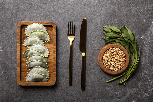 flat lay with served on wooden board green ravioli with flour near bowl with seeds, herb and cutlery