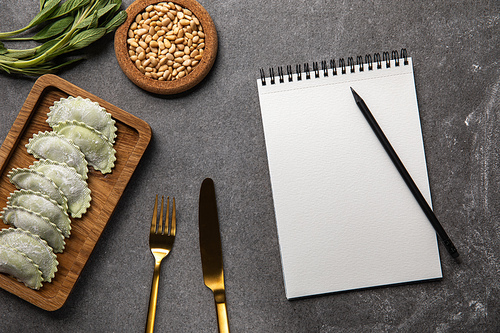 served on wooden board green ravioli with flour near bowl with seeds, herb, cutlery and blank notebook with pencil