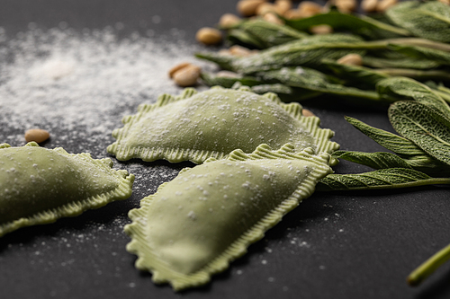 selective focus of green ravioli near sage, flour and pine nuts on black table