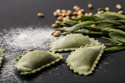 selective focus of green ravioli, sage and scattered flour and pine nuts on black table