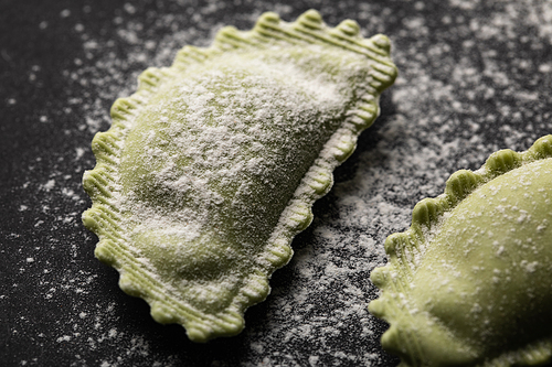 close up view of green fresh ravioli with flour on black surface