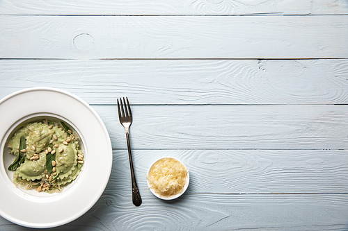 top view of green ravioli served in white plate with pine nuts, sage and grated cheese on white wooden table