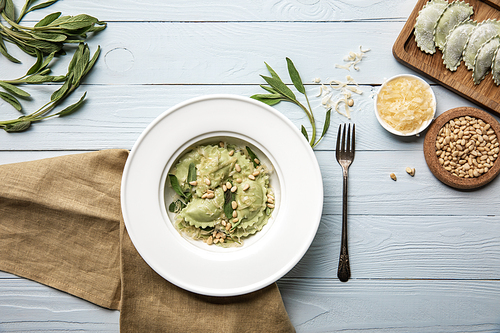 top view of green ravioli served in white plate with pine nuts, sage and grated cheese on white wooden table with fork and sackcloth