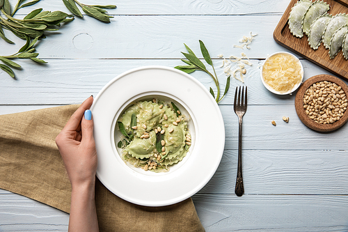 cropped view of woman holding plate with green ravioli, pine nuts, sage near grated cheese on white wooden table with fork and sackcloth