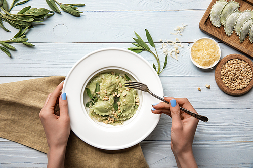 cropped view of woman eating green ravioli with pine nuts and sage near grated cheese on white wooden table