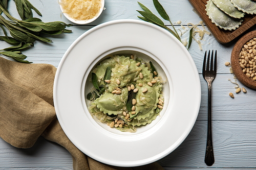 top view of green ravioli served in white plate with pine nuts, sage and grated cheese on white wooden table with fork and napkin