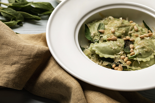 close up view of green ravioli served in white plate with sage, pine nuts and grated cheese near sackcloth