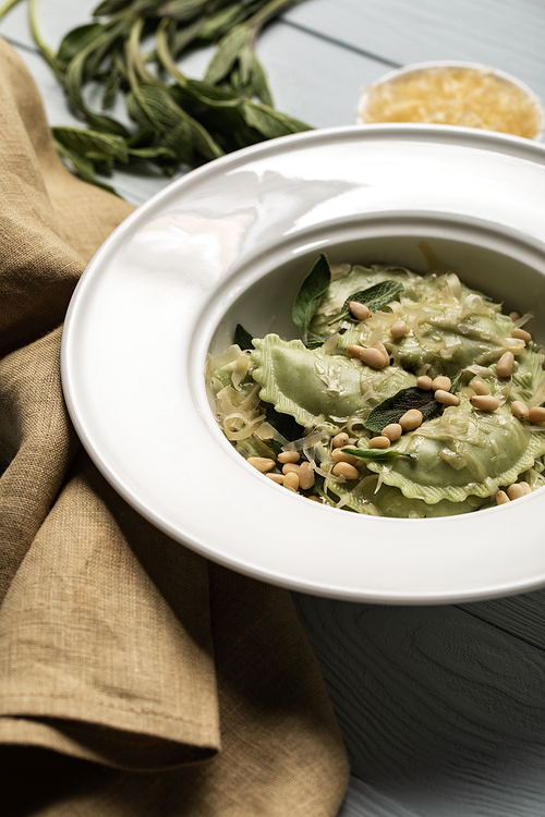 close up view of green ravioli served in white plate with sage, pine nuts and grated cheese near napkin