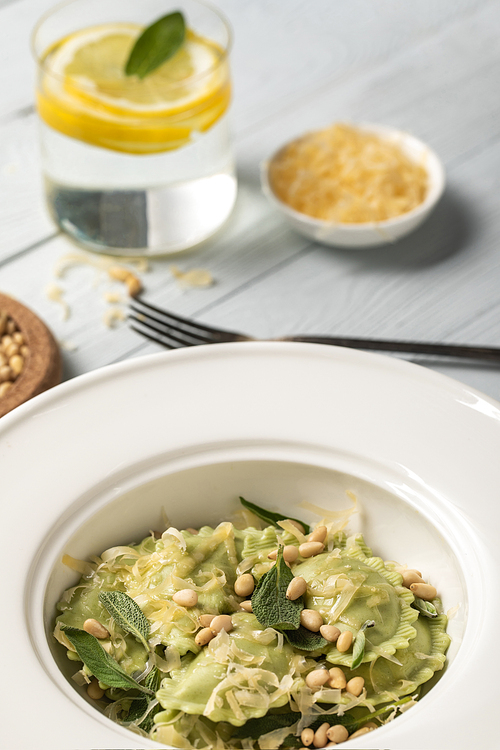 close up view of green ravioli with pine nuts, grated cheese and sage at wooden table near water with lemon and fork