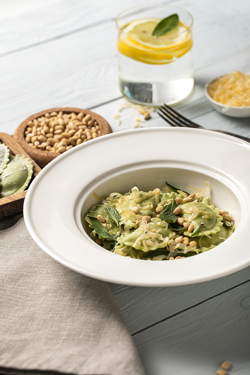 close up view of green ravioli with pine nuts, grated cheese and sage at wooden table near drink with lemon