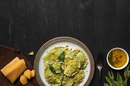 top view of green ravioli served in vintage plate near silver fork, napkin and cheese at black wooden table with copy space