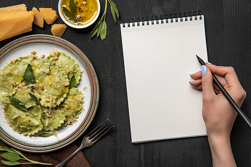 cropped view of woman writing in blank notebook with pencil near served green ravioli