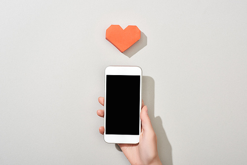 Top view of girl holding smartphone with blank screen by heart shaped paper on grey background