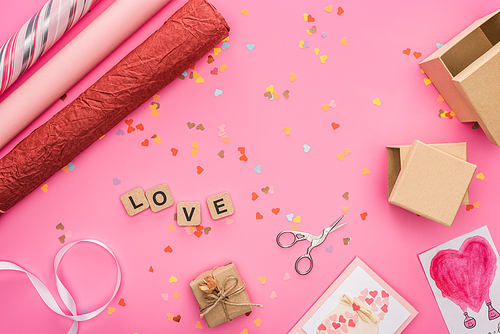 top view of valentines confetti, scissors, wrapping paper, gift boxes, greeting cards and love lettering on wooden cubes on pink background