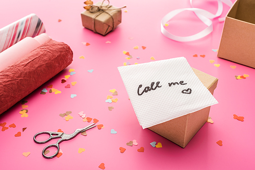 valentines confetti, scissors, card with call me lettering, wrapping paper, gift box on pink background