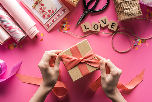 partial view of woman decorating gift box with ribbon near valentines handiwork supplies on pink background