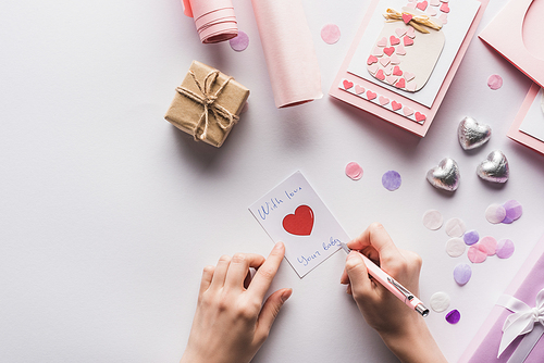 cropped view of woman writing on card near valentines decoration, gifts, hearts and wrapping paper on white background