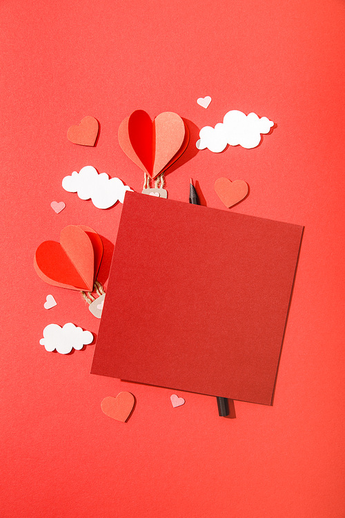 top view of paper heart shaped air balloons in clouds near blank card and pencil on red background