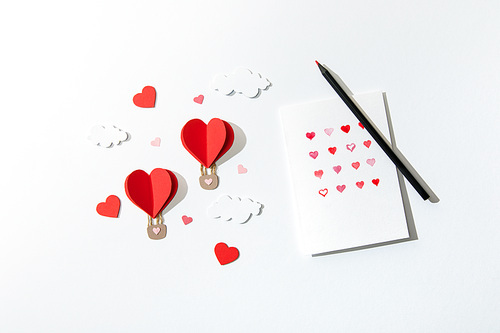 top view of greeting card with hearts and pencil near paper heart shaped air balloons in clouds on white background