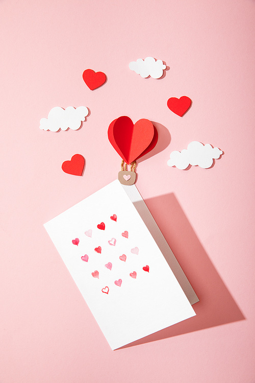 top view of greeting card with hearts in white envelope near paper heart shaped air balloon in clouds on pink