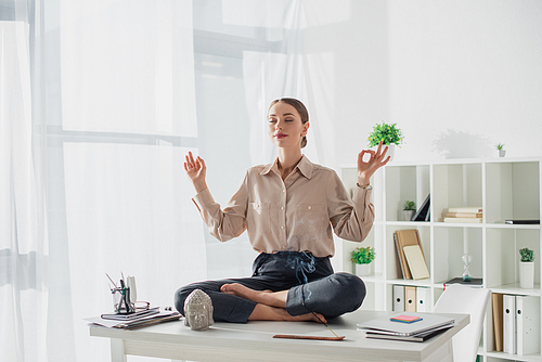 attractive businesswoman meditating in lotus pose with gyan mudra at workplace with Buddha head and incense stick