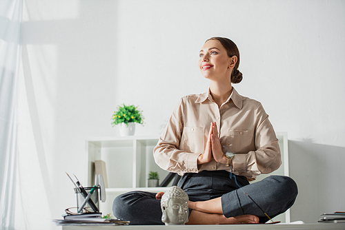 happy businesswoman meditating in lotus pose with namaste gesture at workplace with Buddha head and incense stick