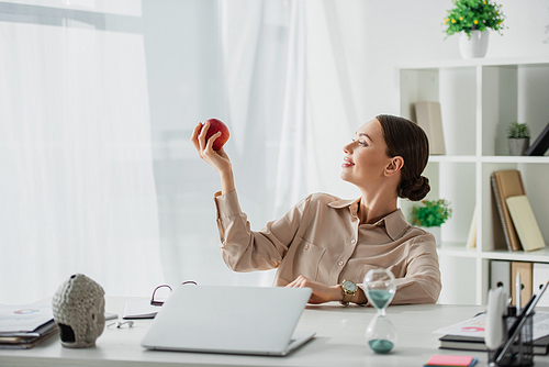 beautiful smiling businesswoman holding apple at workplace with Buddha head, laptop and sand clock in office