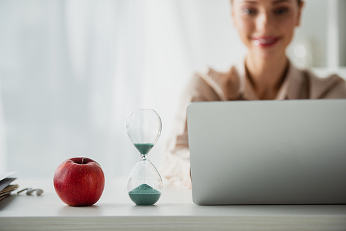 selective focus of smiling businesswoman sitting at workplace with apple, sand clock and laptop in office