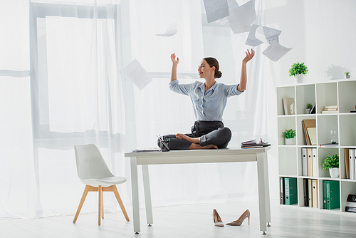 happy businesswoman sitting in lotus pose on table with falling business papers in office
