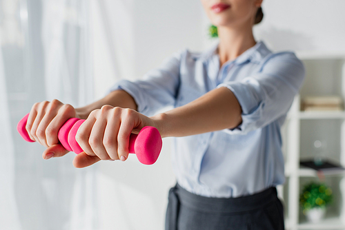 cropped view of businesswoman training with pink dumbbells in office, selective focus