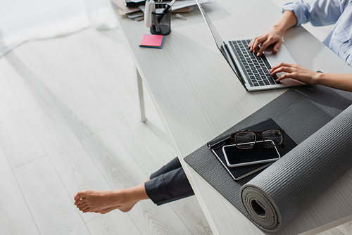 cropped view of barefoot businesswoman working on laptop at workplace with yoga mat, notepad, smartphone and eyeglasses