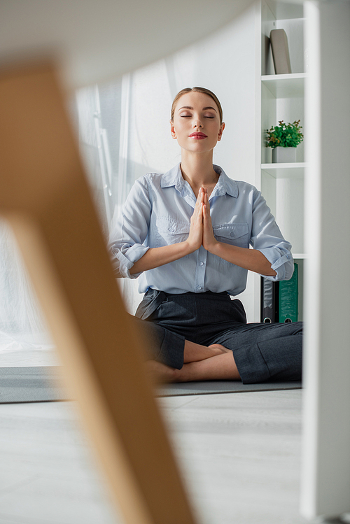 selective focus of businesswoman practicing yoga in lotus position with namaste gesture on mat in office